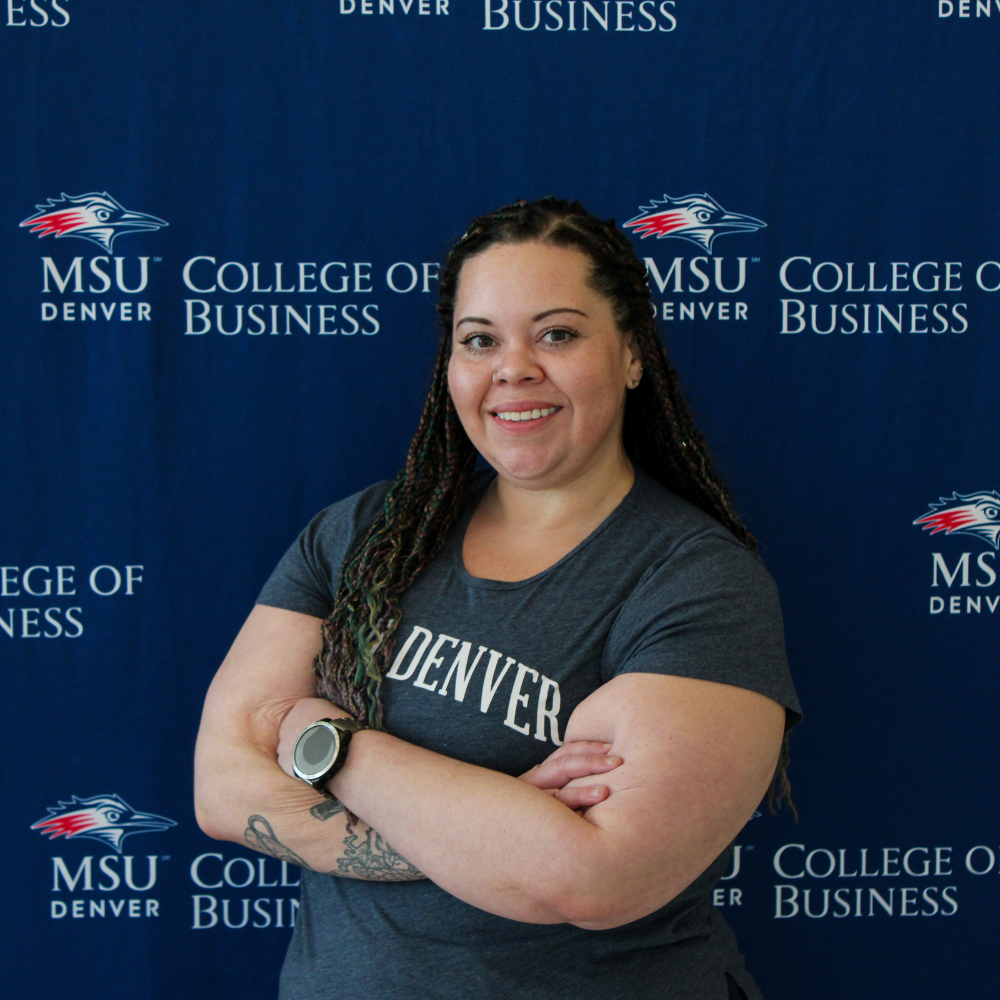 A smiling student ambassador with long braids and crossed arms, wearing a grey MSU Denver t-shirt in front of a College of Business backdrop.