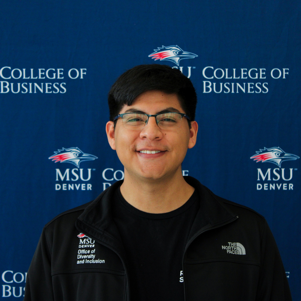 A smiling student ambassador wearing glasses and a black MSU Denver Office of Diversity and Inclusion jacket in front of a College of Business backdrop.