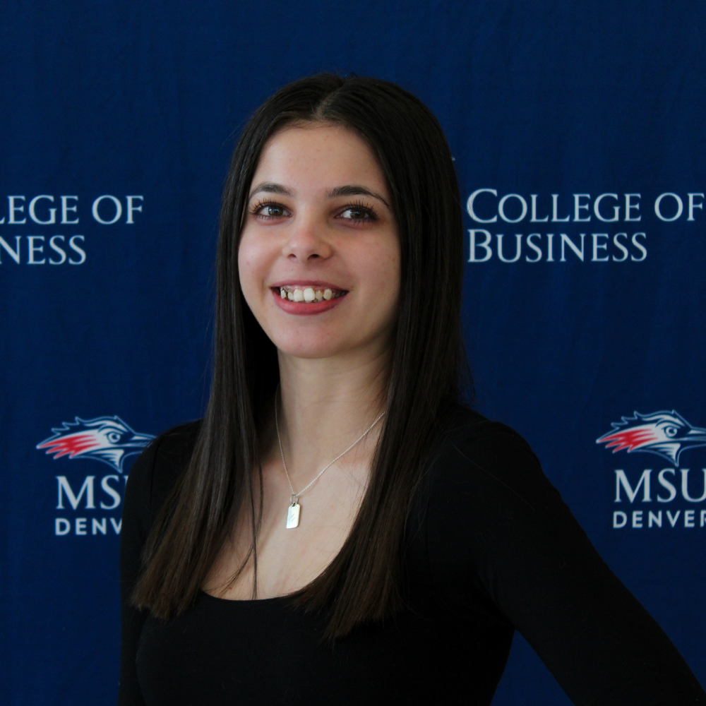 A smiling student ambassador with long, dark straight hair wearing a black top and a silver necklace in front of an MSU Denver College of Business backdrop.