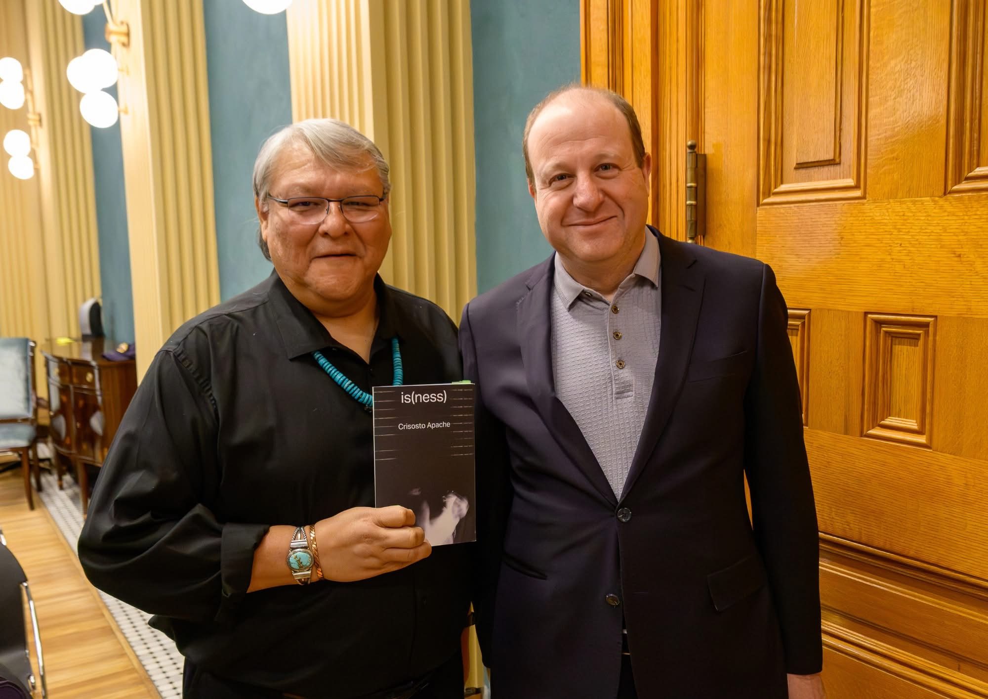 Crisosto Apache, Colorado Poet Laureate, holding a copy of the poetry collection is(ness) with Governor Jared Polis.