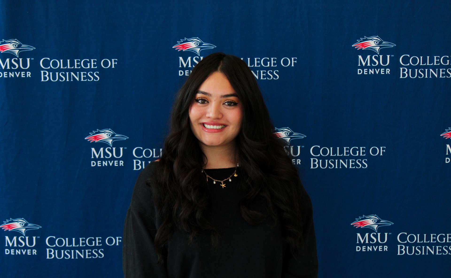 A smiling student ambassador with long, dark wavy hair, wearing a black top and a gold necklace with charms in front of an MSU Denver College of Business backdrop.
