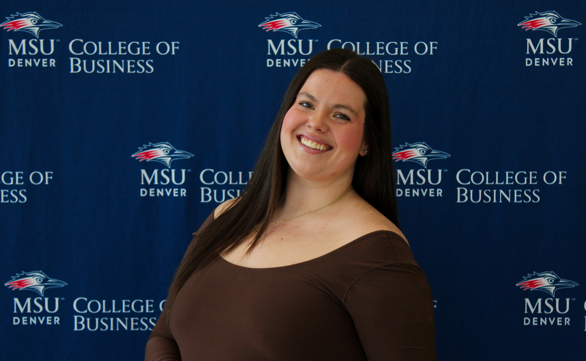 A smiling student ambassador with long, dark wavy hair, wearing a black top and a gold necklace with charms in front of an MSU Denver College of Business backdrop.