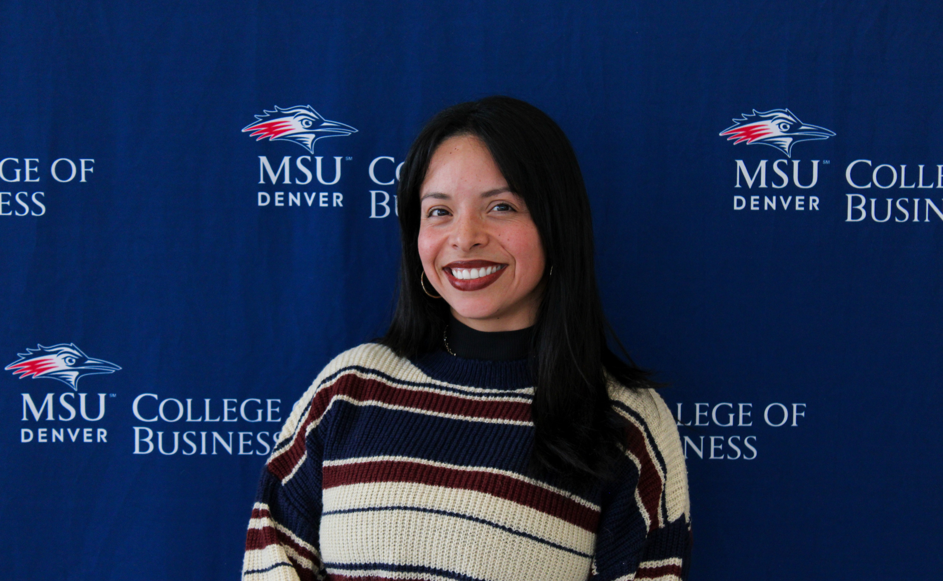 A smiling student ambassador with long, dark wavy hair, wearing a black top and a gold necklace with charms in front of an MSU Denver College of Business backdrop.
