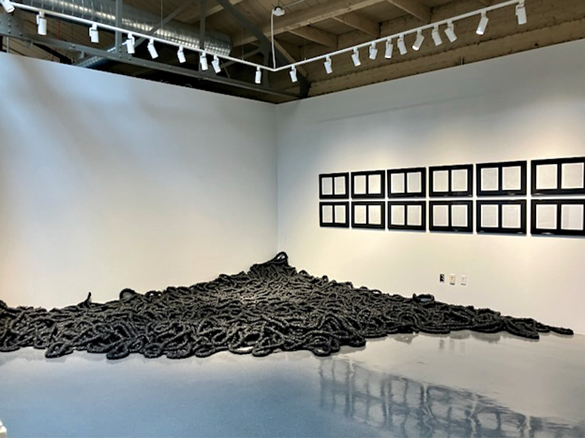 A gallery space with a black rope-like installation in the corner and framed documents on the wall.