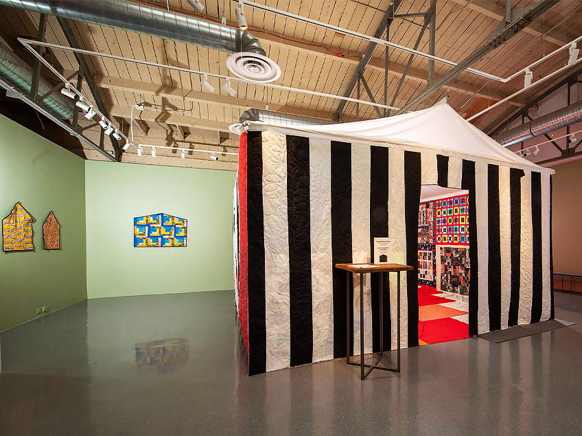 Art gallery with a black and white striped tent structure and quilts displayed.