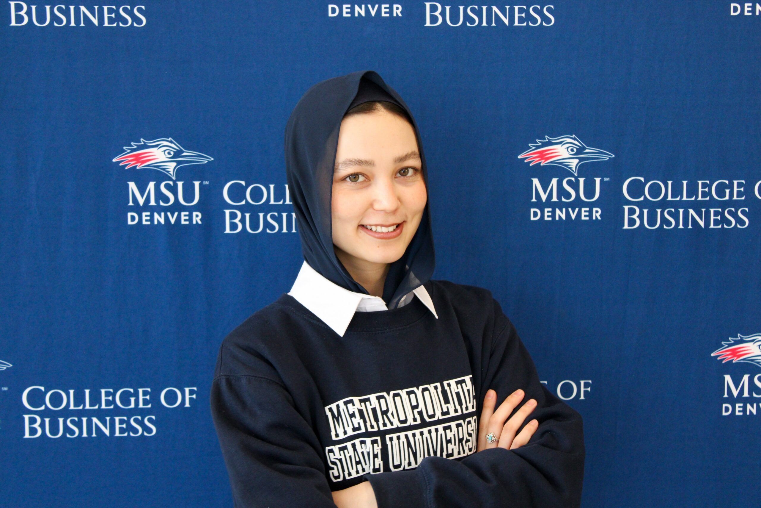 A headshot of Madina smiling with her arms crossed, wearing a navy blue Metropolitan State University sweatshirt over a white collared shirt and a dark blue hijab. She is standing in front of a blue MSU Denver College of Business backdrop.