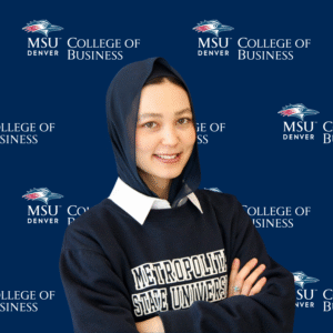 A headshot of Madina smiling with her arms crossed, wearing a navy blue Metropolitan State University sweatshirt over a white collared shirt and a dark blue hijab. She is standing in front of a blue MSU Denver College of Business backdrop.
