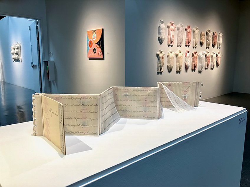 An accordion-style book on a pedestal in an art gallery with various artworks on the walls.