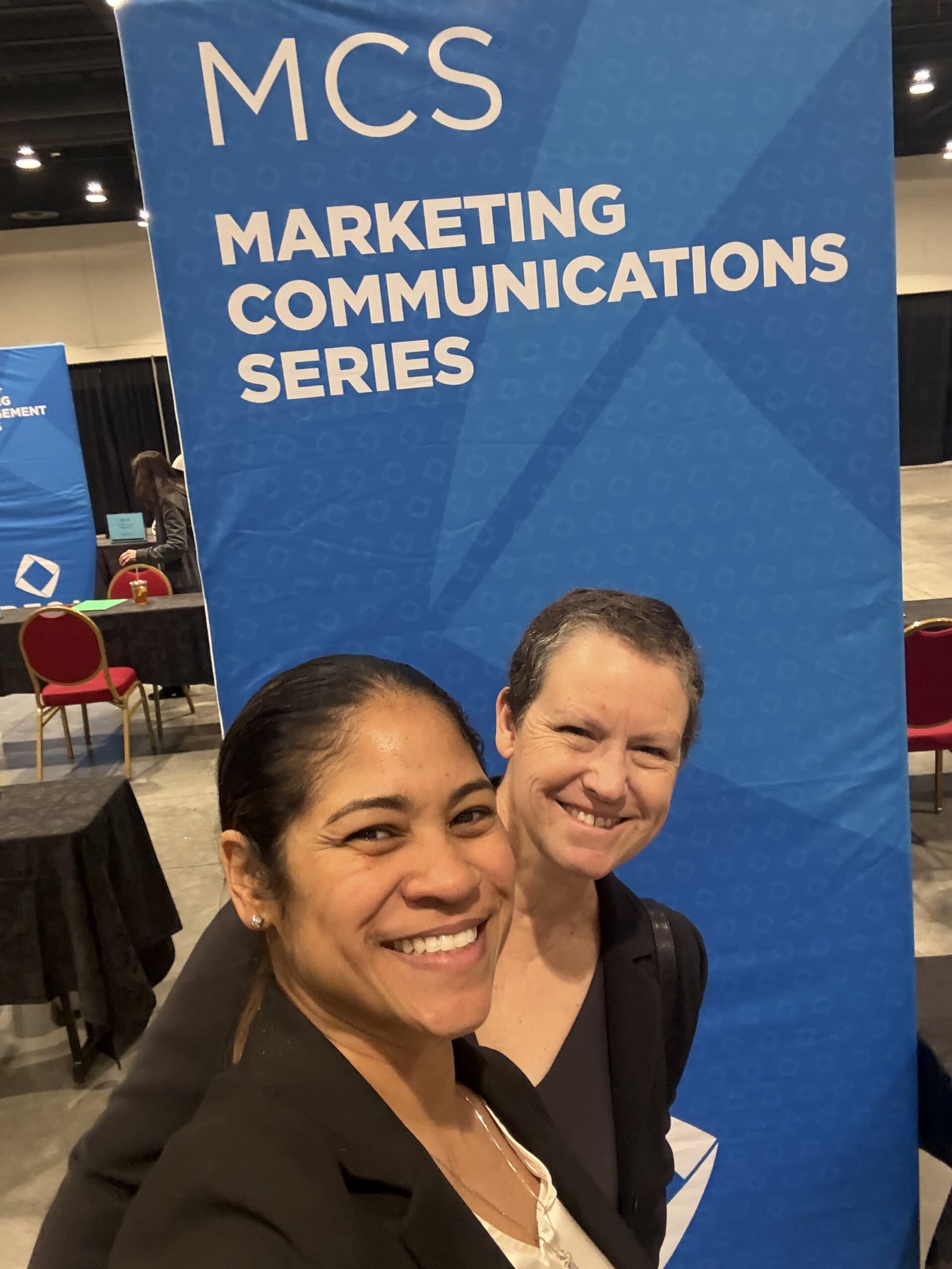 Two individuals taking a photo in front of a large blue banner that reads ‘MCS Marketing Communications Series’ at an indoor event with tables and chairs in the background.