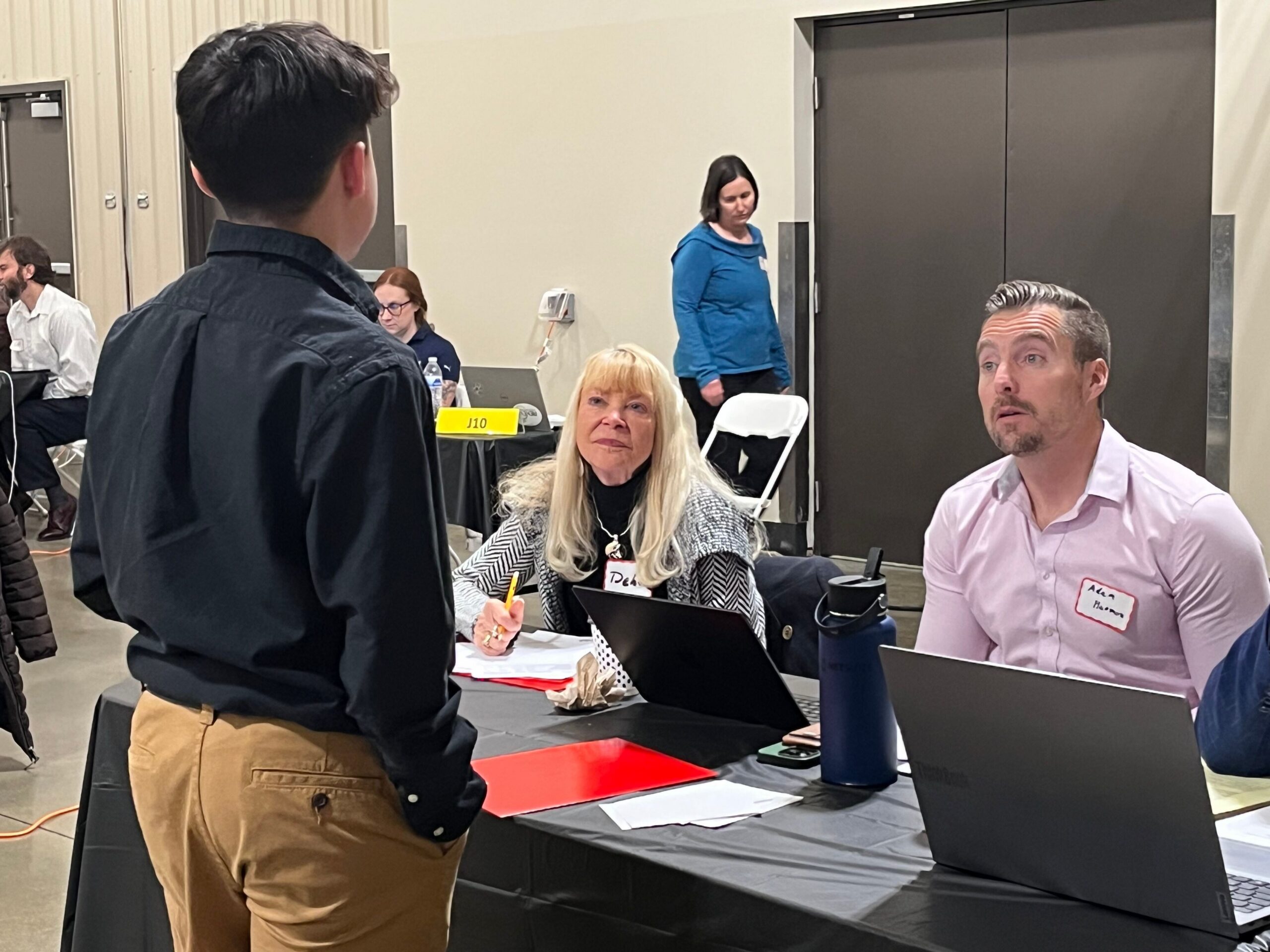 A student in a black button-down shirt and khaki pants stands with their back to the camera, speaking to two seated professionals tabling for the FBLA event.