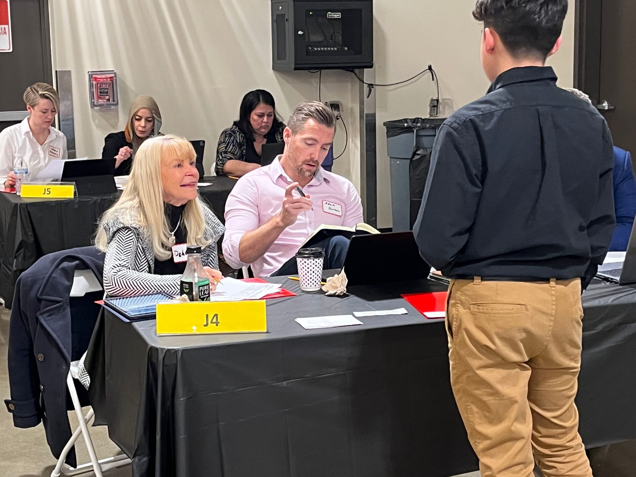 A student in a black button-down shirt and khaki pants stands with their back to the camera, speaking to two seated professionals tabling for the FBLA event.