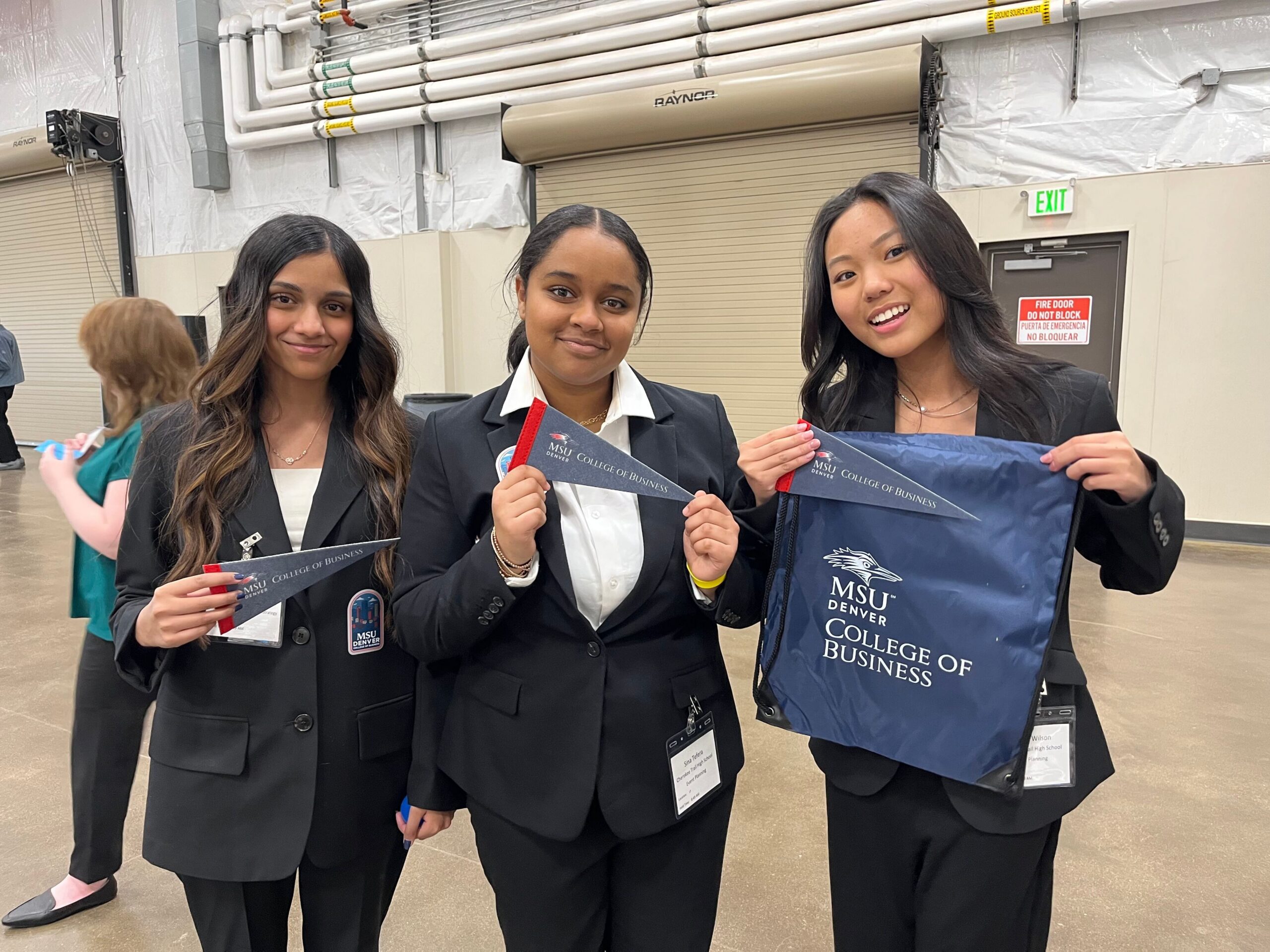 Three young women in matching black professional blazers pose for a photo. The student on the left and the student in the middle hold small navy blue pennants that read 