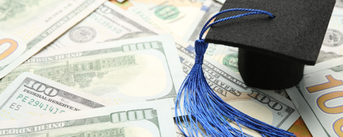 A small black graduation cap with a long blue tassel sits atop a scattered pile of United States one-hundred-dollar bills.