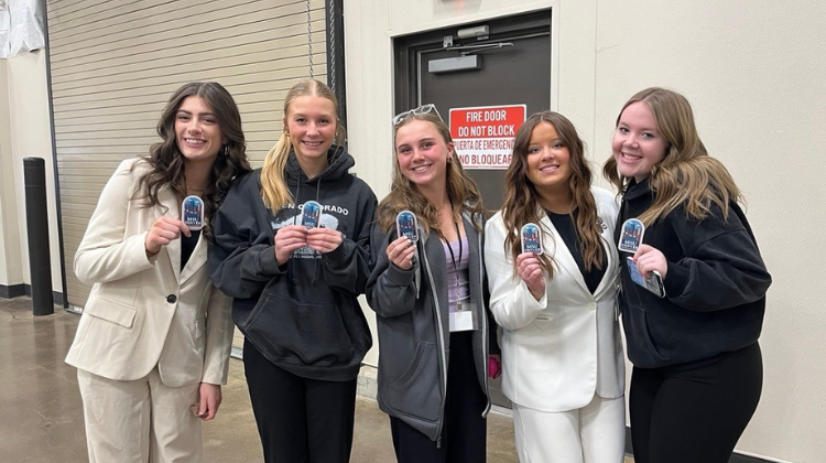 Five smiling young women stand together in an indoor facility, posing for a photo. They are holding up small, blue MSU Denver stickers. Two of the women are wearing professional cream-colored suits, while the others are dressed in casual dark hoodies and jackets. An exit door with a red 