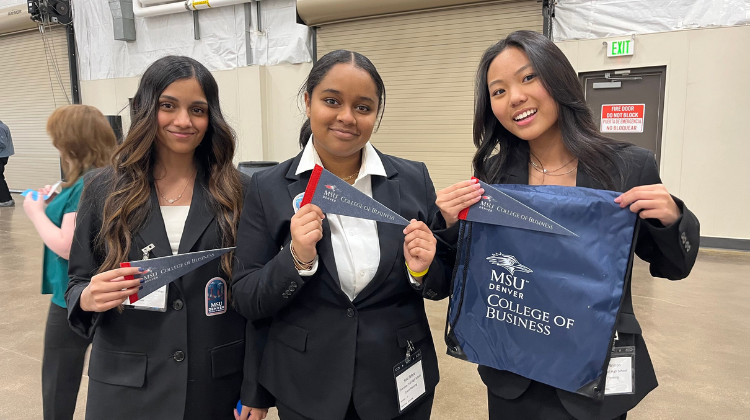 Three young women in matching black professional blazers pose for a photo. The student on the left and the student in the middle hold small navy blue pennants that read 