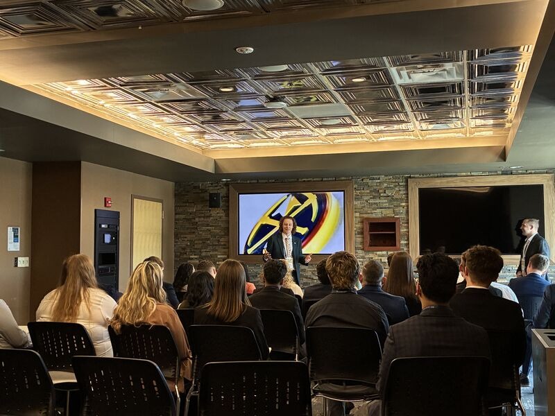 A speaker presenting to an audience seated in a conference room with a screen at the front.