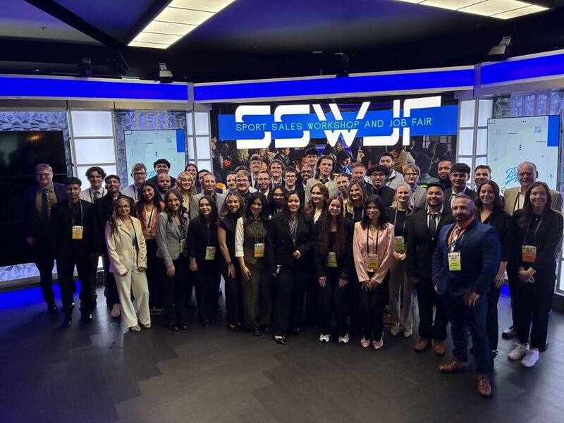 Gemini said A group of over 40 young professionals and attendees stand together for a large group portrait inside a modern, brightly lit event space with blue accent lighting. The background features a large digital screen with the bold text 