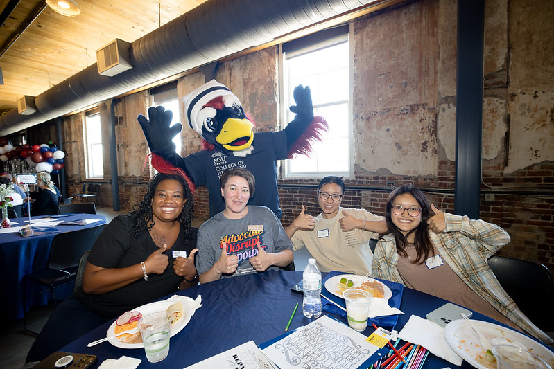 Four people sitting at a table with Rowdy the Mascot behind them. Everyone shows a thumbs up and smiles!