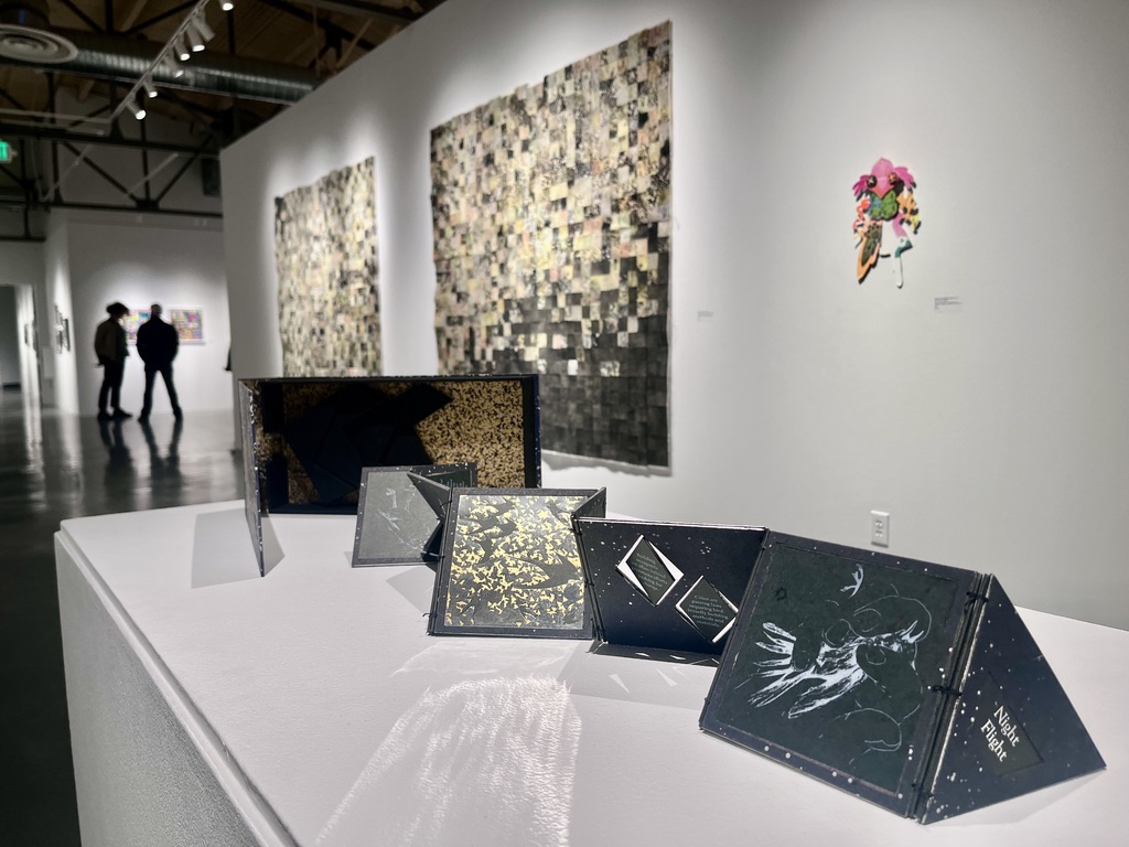 A handprinted artist book on a pedestal with more works of printmaking on the wall behind.