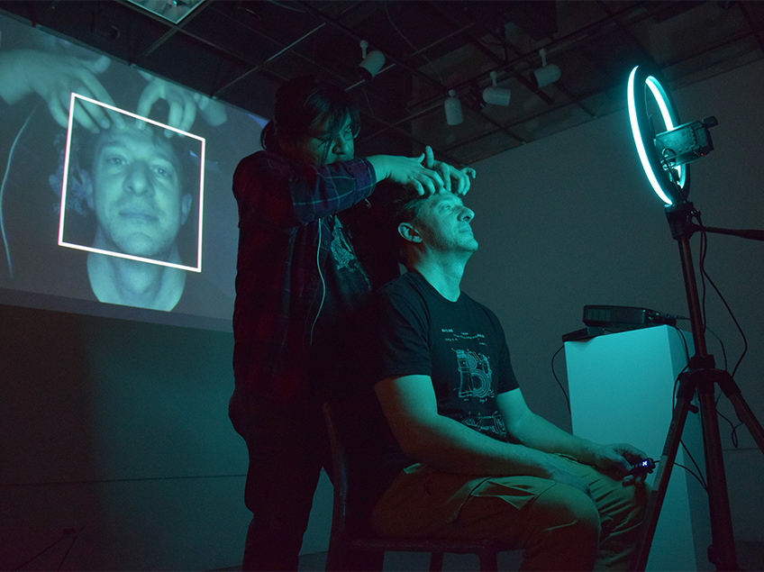A live capture of a student manipulating another student's face is projected onscreen behind them.