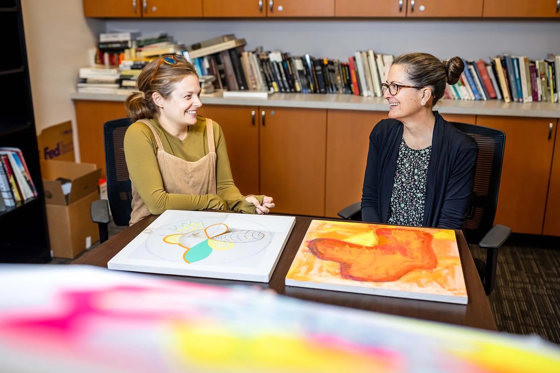 Two women sit in front of colorful canvases