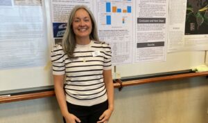 A woman with gray hair and a striped shirt standing in front of a research-focused poster.