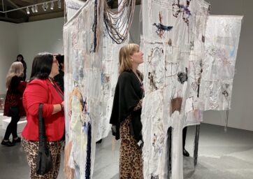 People observe a textile art installation with detailed stitching and colorful threads in a gallery.