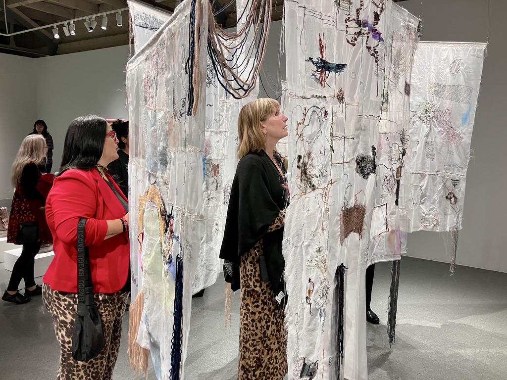 People observe a textile art installation with detailed stitching and colorful threads in a gallery.