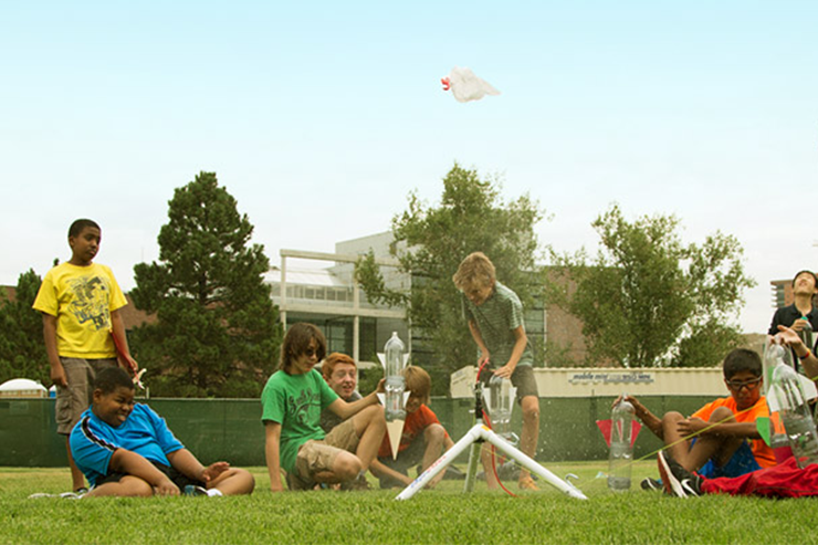 Children on a campus lawn gather around a boy launching a toy rocket
