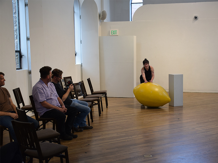 Student pushes enormous waist-high lemon in Emmanuel Gallery while professors look on.