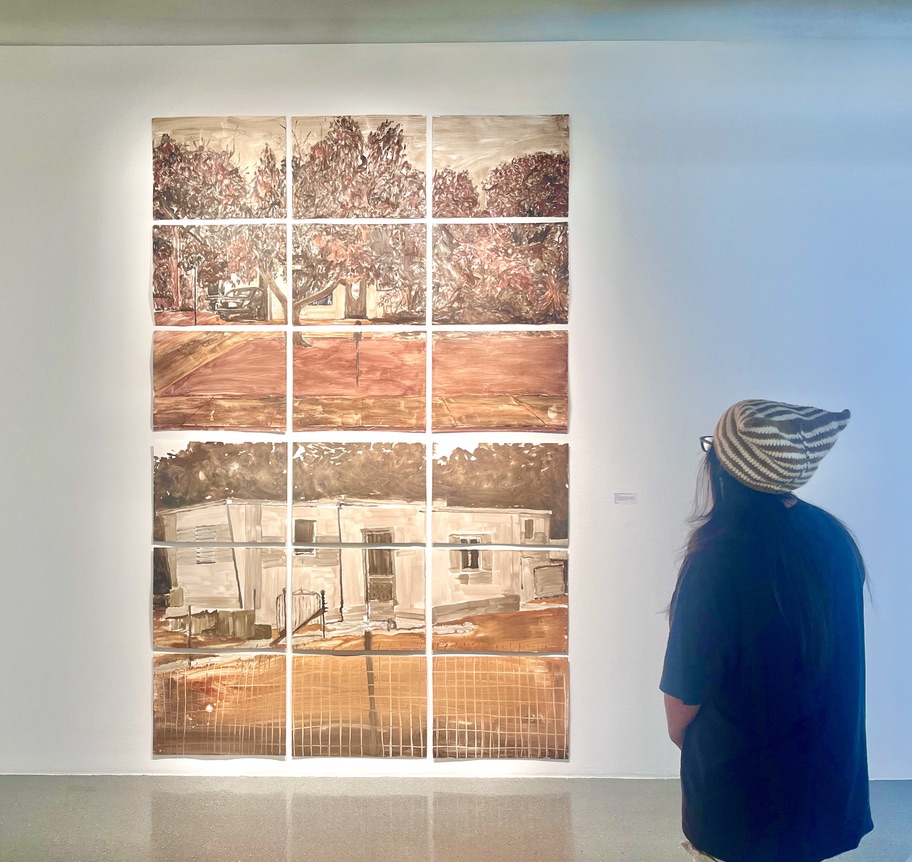 A person stands in front of a gallery wall that displays two large grids of 9 monotype prints. Each grid forms a sepia-toned image, one of a house surrounded by trees and the other of a mobile home.