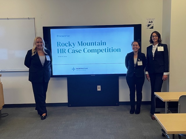 The three team members pose together in front of their introductory presentation slide in a classroom setting. All three women are wearing dark business suits and name tags for the competition.