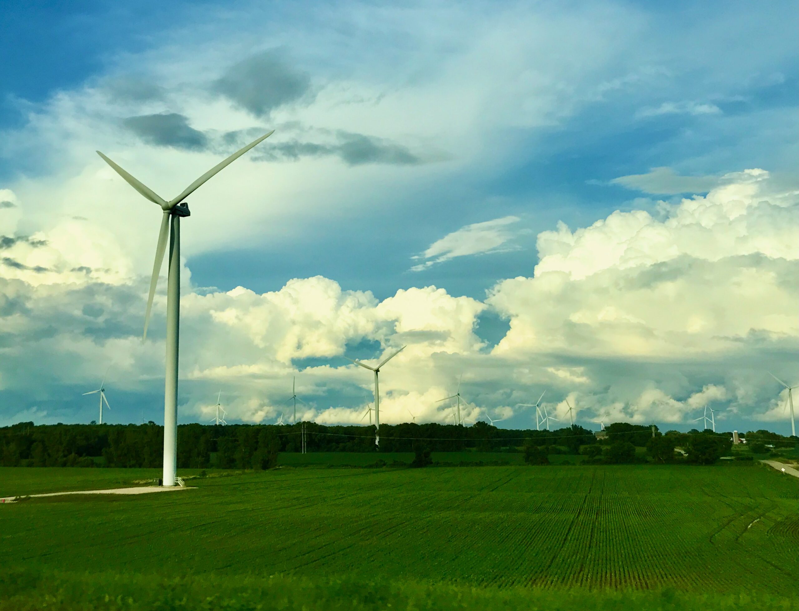 Photograph of several wind turbines in a green field with white puffy clouds.