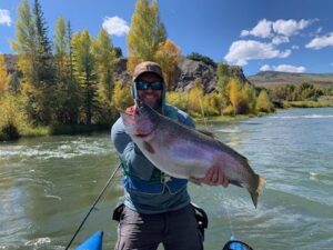 Man with a hat, beard and sunglasses holds a fish in a river surrounded by trees.