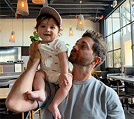 Man in a tshirt holds a baby wearing a hat on his shoulders
