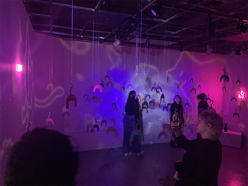 Many copies of a pattern with u-shape dangle from ceiling in a dark and moodily lit gallery while observers walk through the space.