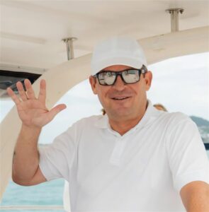 Man with a white shirt and white hat on waves at the camera,