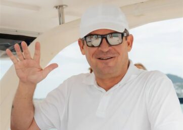 Man with a white shirt and white hat on waves at the camera,