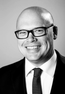 Black and white photo of man wearing glasses and wearing a white shirt, polka-dot tie, and black jacket