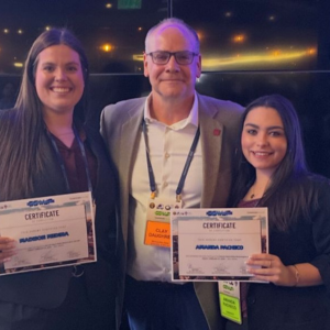 Three people stand together and smile, holding certificates. Clay Daugherty is in the center, flanked by Madison Redina and Amanda Pacheco, who are holding their certificates. They are all wearing event lanyards and business attire in front of a dark backdrop.