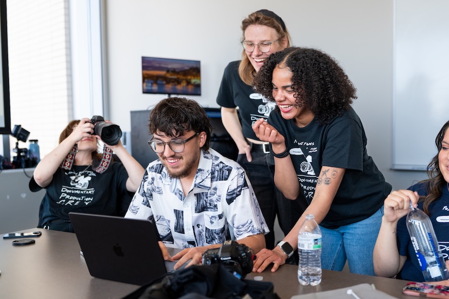 A group of students gathered around a laptop. One student holds a camera.