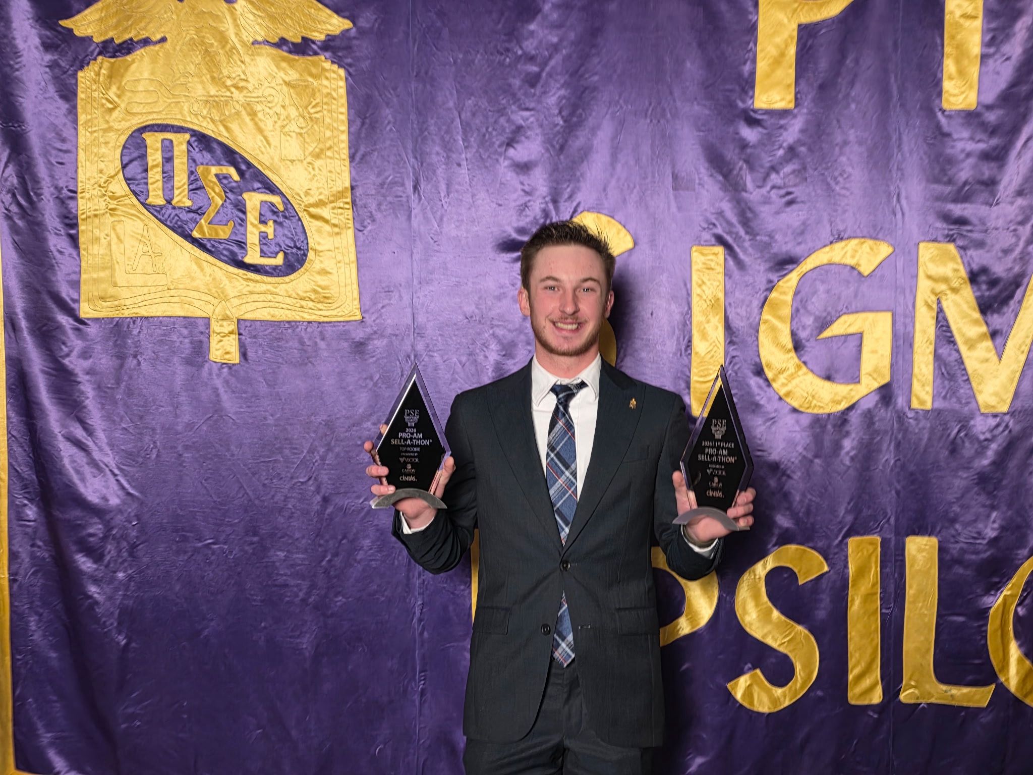Nick Russo in a dark suit and blue patterned tie stands smiling in front of a purple and gold Pi Sigma Epsilon (PSE) banner. He is holding two glass trophies; the one in his left hand reads 