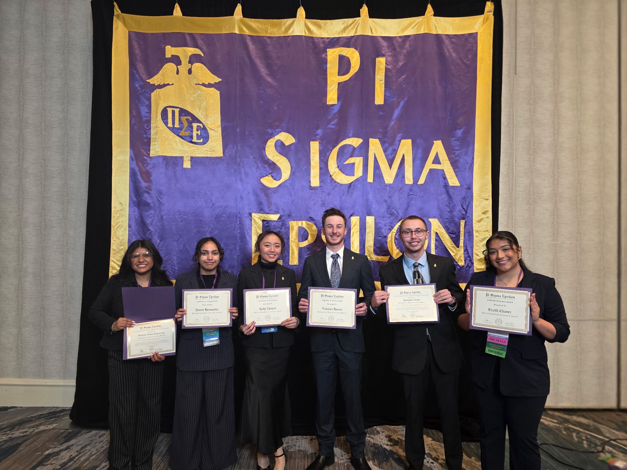 Six PSE students stand in a row in front of a large purple PSE banner, each holding a framed award certificate. They are dressed in professional business attire and are smiling at the camera in a brightly lit conference room.