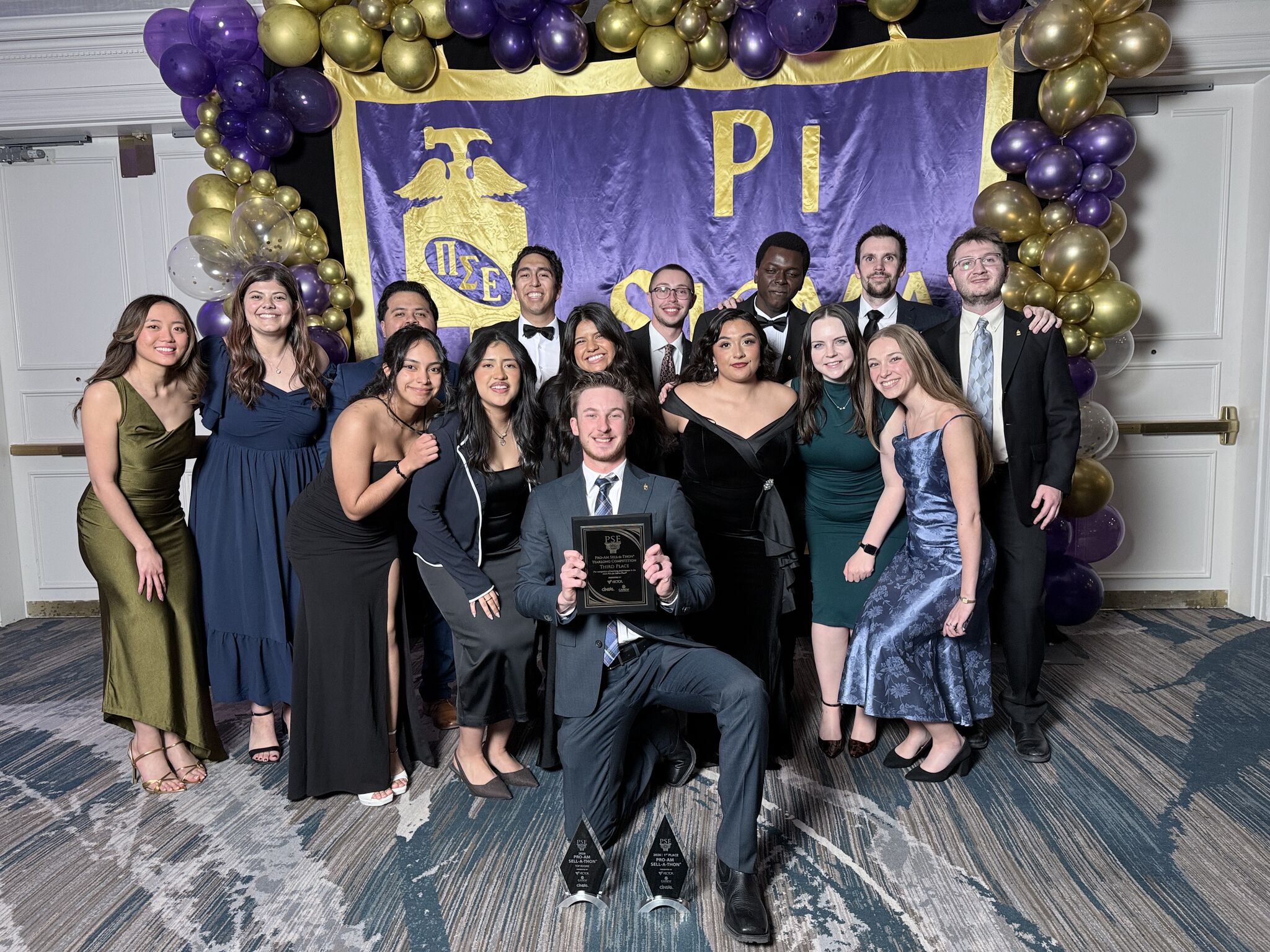 A group of about 15 students in formal evening wear pose for a group photo in front of a purple banner decorated with a purple and gold balloon arch. In the center, a young man is kneeling and holding a black award plaque, with two glass trophies placed on the floor in front of him.