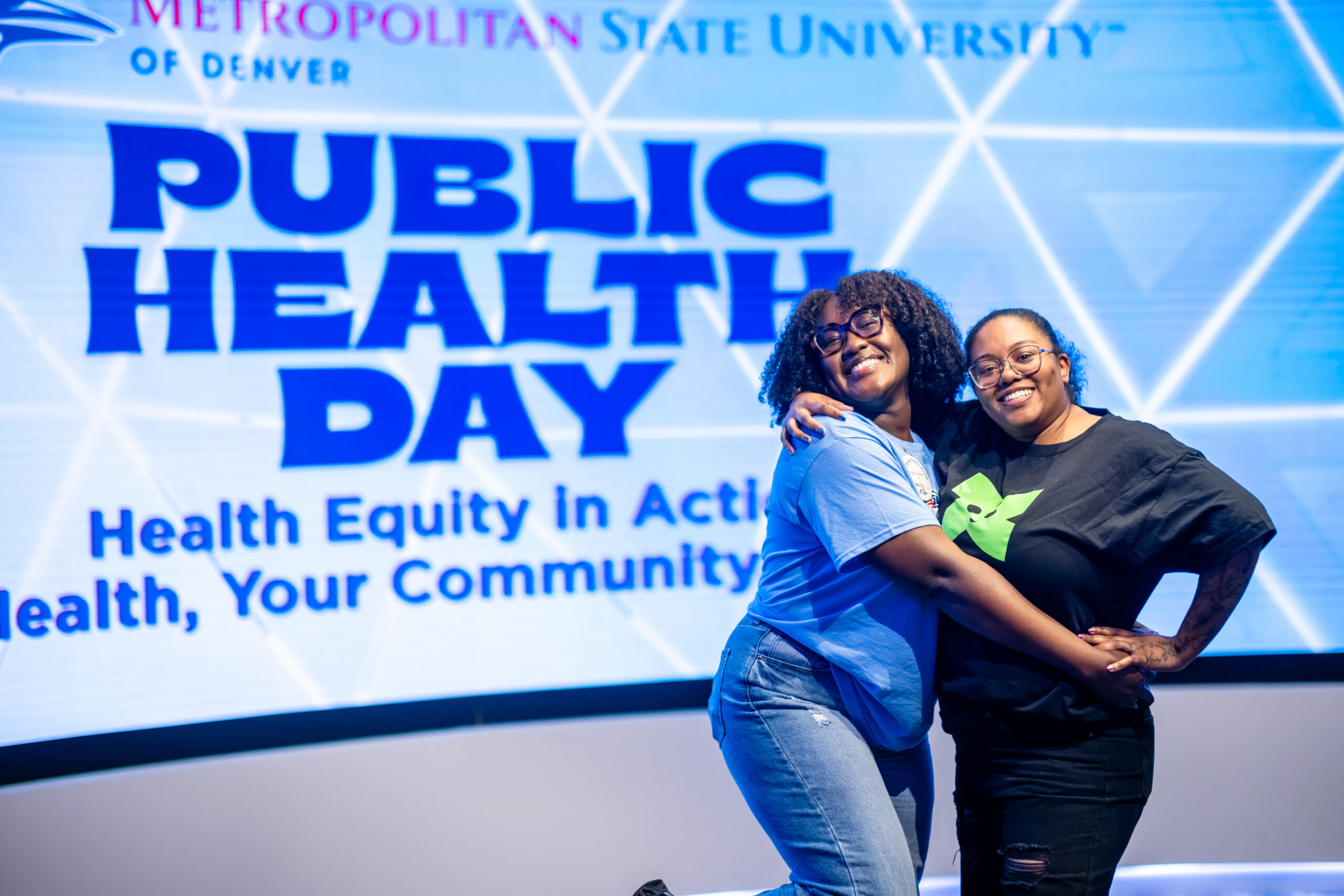 Two woman hugging and smiling with the words Public Health Day on a screen in the background