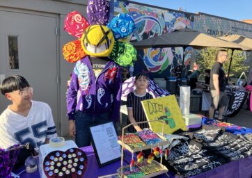 A table covered in small items such as buttons, keychains, and figurines. Two young people sit behind the table and a third stands while wearing a rainbow colored, cartoon flower mask over his head.