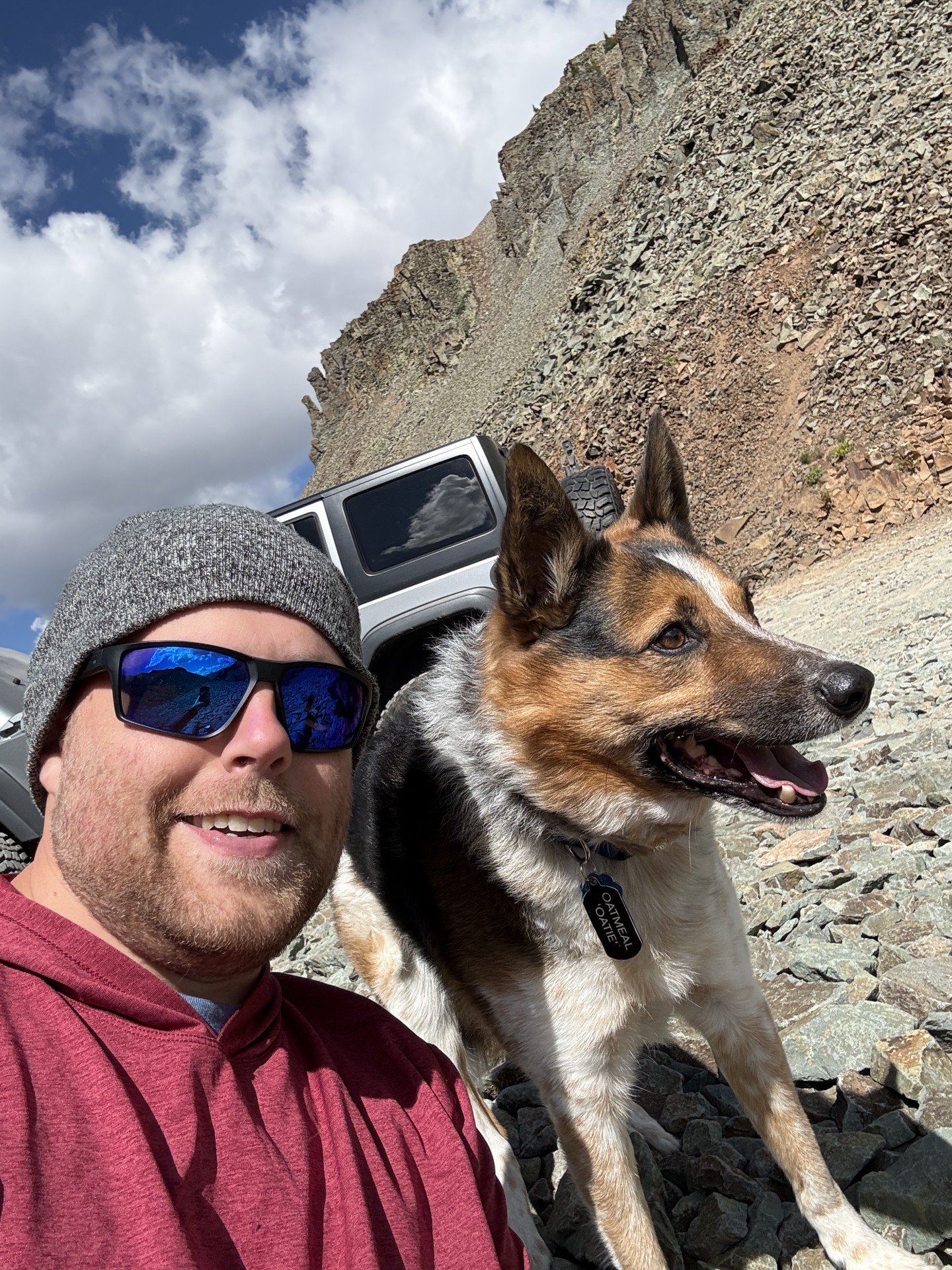 Man wearing sunglasses posing next to a tri-color dog, with a rocky terrain background.