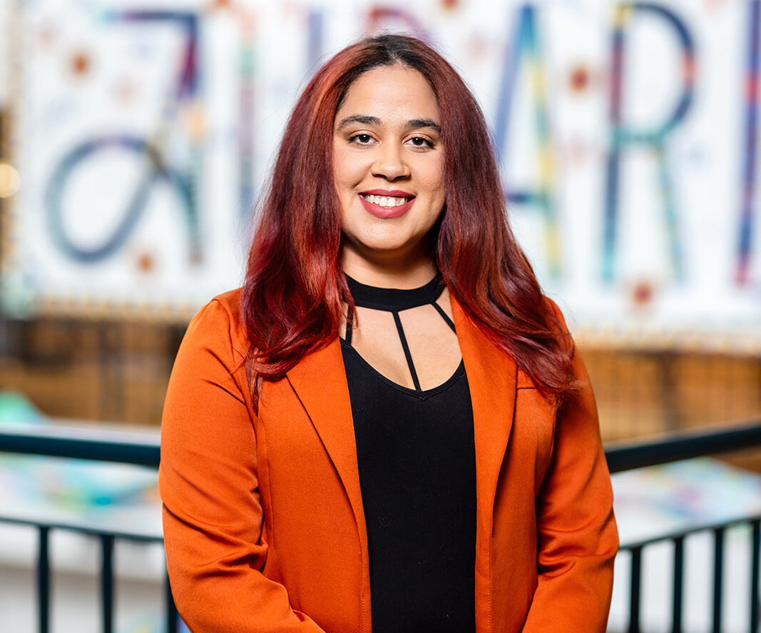 Female student wearing a bright orange jacket poses in front of an Auraria sign