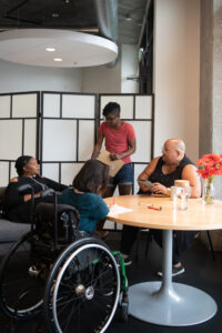 Four disabled people of color gather around a table during a meeting. A Black woman sitting on a couch speaks with a neutral expression while the three others (a South Asian person sitting in a wheelchair and taking notes, a Black non-binary person sitting in a chair with a tablet and cane, and a Black non-binary person standing with a clipboard) listen.