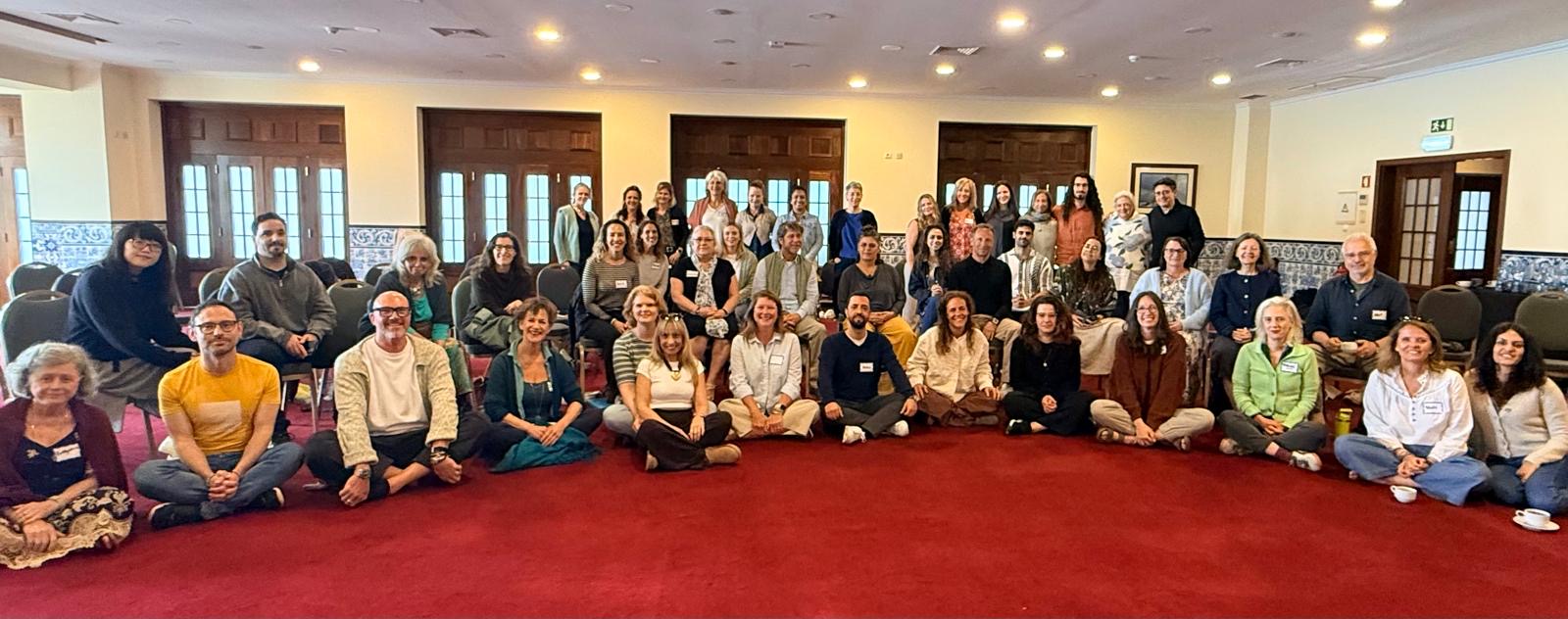 A large group of adults seated and standing in rows inside a carpeted conference room with wooden doors and patterned wall tiles, facing the camera.